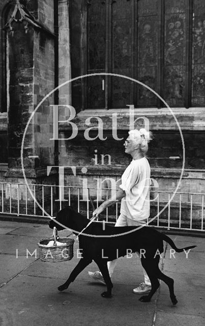 A woman walking her dog with a basket in its mouth outside Bath Abbey c.1980